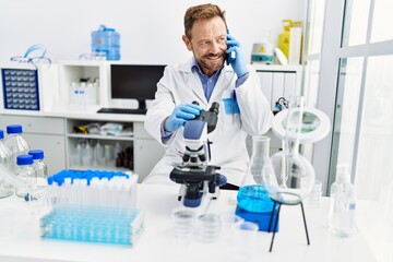 Middle age hispanic man wearing scientist uniform talking on the smartphone using microscope at laboratory