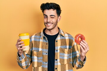 Young hispanic man eating doughnut and drinking coffee winking looking at the camera with sexy...