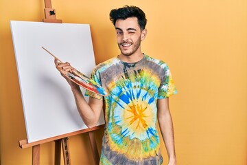 Young hispanic man standing drawing with palette by painter easel stand winking looking at the...