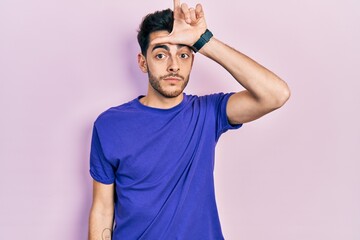 Young hispanic man wearing casual t shirt making fun of people with fingers on forehead doing loser...