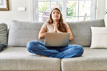 Beautiful young brunette woman sitting on the sofa using computer laptop at home begging and...