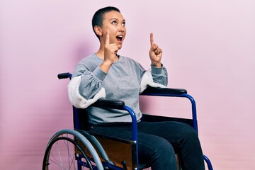 Beautiful hispanic woman with short hair sitting on wheelchair amazed and surprised looking up and...