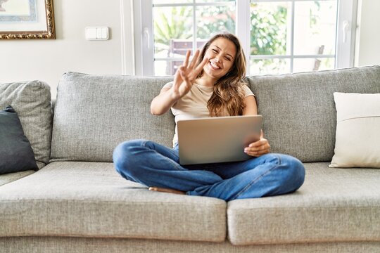 Beautiful young brunette woman sitting on the sofa using computer laptop at home showing and pointing up with fingers number three while smiling confident and happy.