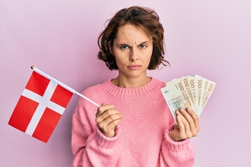 Young brunette woman holding norway flag and krone banknotes skeptic and nervous, frowning upset...