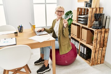 Beautiful caucasian business woman working at the office sitting on pilates ball smiling happy pointing with hand and finger to the side