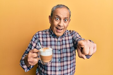 Handsome senior man with grey hair drinking a cup coffee pointing to you and the camera with fingers, smiling positive and cheerful