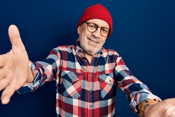 Handsome mature man wearing hipster look with wool cap looking at the camera smiling with open arms for hug. cheerful expression embracing happiness.