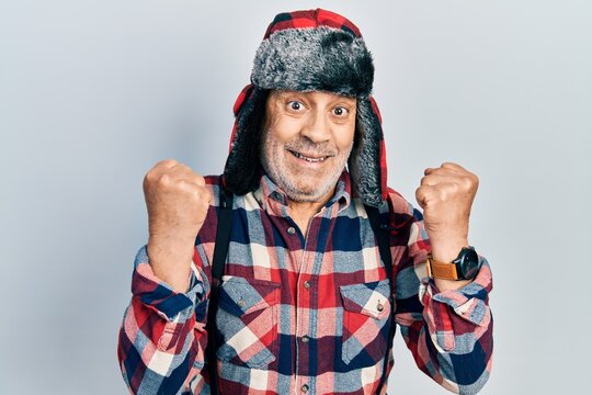 Handsome mature man wearing winter hat with ear flaps screaming proud, celebrating victory and success very excited with raised arms