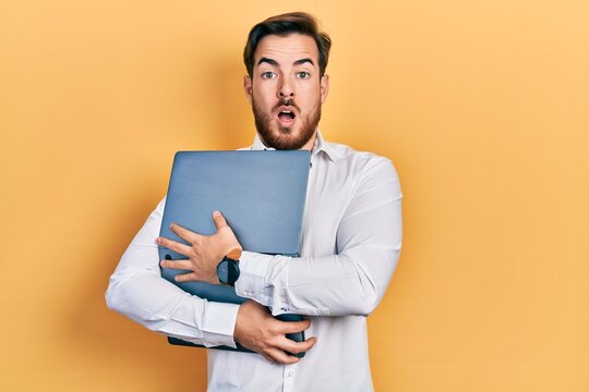 Handsome caucasian man with beard hugging laptop afraid and shocked with surprise and amazed expression, fear and excited face.