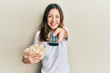 Young brunette woman eating popcorn using tv control smiling with a happy and cool smile on face....