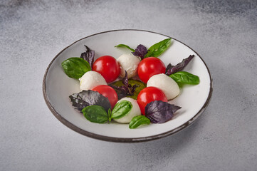 Delicious italian caprese salad with ripe tomatoes, fresh basil and mozzarella cheese in white plate on grey background. Close up 
