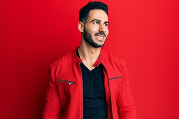 Obraz premium Young hispanic man wearing red leather jacket looking away to side with smile on face, natural expression. laughing confident.