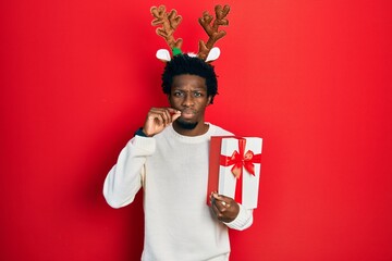 Young african american man wearing deer christmas hat holding gift mouth and lips shut as zip with fingers. secret and silent, taboo talking