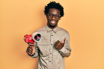 Young african american man holding packing tape machine smiling happy and positive, thumb up doing...