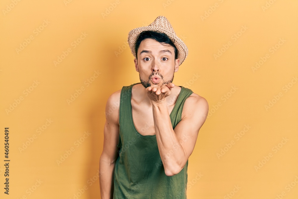 Poster young hispanic man wearing summer hat looking at the camera blowing a kiss with hand on air being lo