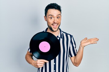 Young hispanic man holding vinyl disc celebrating achievement with happy smile and winner expression with raised hand
