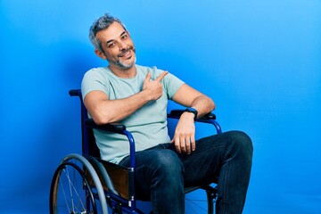 Handsome middle age man with grey hair sitting on wheelchair cheerful with a smile on face pointing with hand and finger up to the side with happy and natural expression