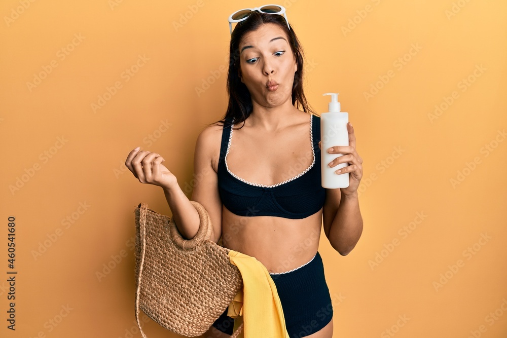 Poster young latin woman wearing bikini and hat holding summer wicker handbag and sunscreen making fish fac