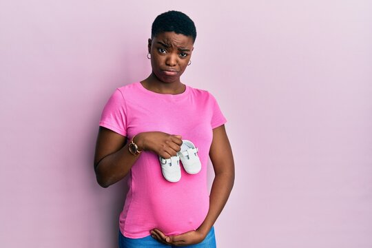 Young african american woman expecting a baby holding shoes skeptic and nervous, frowning upset because of problem. negative person.