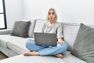 Young caucasian woman using laptop at home sitting on the sofa puffing cheeks with funny face. mouth inflated with air, crazy expression.