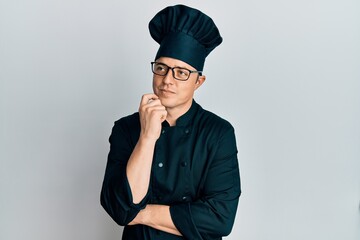 Handsome young man wearing professional cook uniform and hat thinking concentrated about doubt with finger on chin and looking up wondering