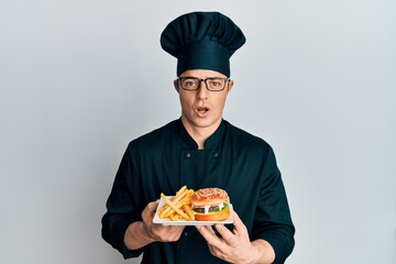 Handsome young man chef holding burger with fries afraid and shocked with surprise and amazed expression, fear and excited face.