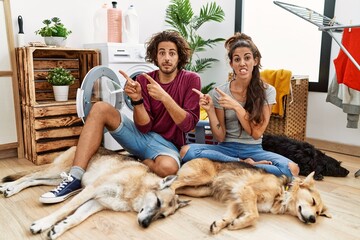 Young hispanic couple doing laundry with dogs pointing aside worried and nervous with both hands, concerned and surprised expression