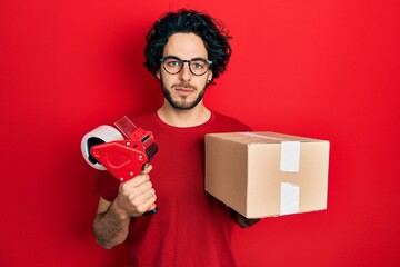 Handsome hispanic man holding packing tape and cardboard box relaxed with serious expression on...