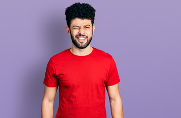 Young arab man with beard wearing casual red t shirt winking looking at the camera with sexy expression, cheerful and happy face.