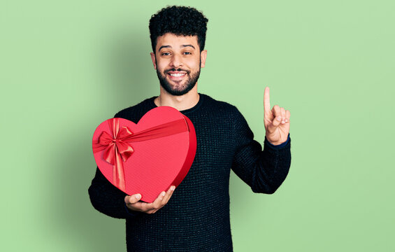 Young arab man with beard holding valentine gift smiling with an idea or question pointing finger with happy face, number one