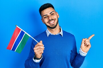 Young hispanic man with beard holding gambia flag smiling happy pointing with hand and finger to...