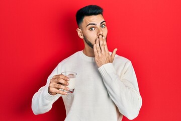 Young hispanic man with beard holding glass of milk covering mouth with hand, shocked and afraid...
