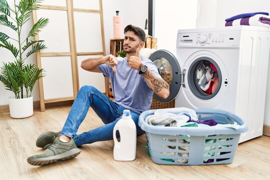 Young hispanic man putting dirty laundry into washing machine doing thumbs up and down, disagreement and agreement expression. crazy conflict