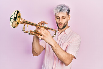 Young hispanic man with modern dyed hair playing trumpet making fish face with mouth and squinting...