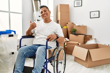Young hispanic man sitting on wheelchair at new house smiling happy and positive, thumb up doing excellent and approval sign