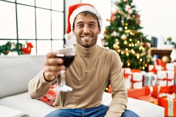 Young hispanic man drinking wine sitting by christmas tree at home