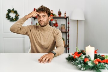 Young handsome man with beard sitting on the table by christmas decoration pointing unhappy to...