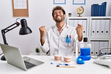 Young hispanic dentist man working at medical clinic very happy and excited doing winner gesture with arms raised, smiling and screaming for success. celebration concept.