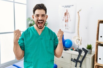 Young man with beard working at pain recovery clinic celebrating surprised and amazed for success with arms raised and open eyes. winner concept.