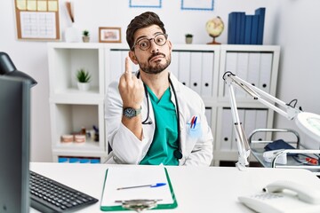Young man with beard wearing doctor uniform and stethoscope at the clinic showing middle finger,...