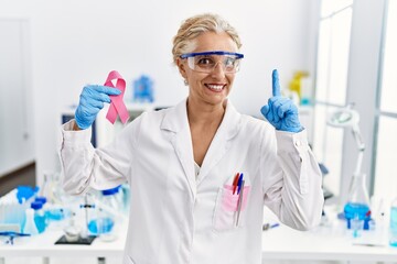 Middle age blonde woman working at laboratory looking for breast cancer cure surprised with an idea or question pointing finger with happy face, number one