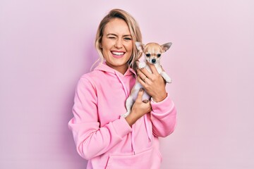 Beautiful young blonde woman hugging cute chihuahua dog winking looking at the camera with sexy...