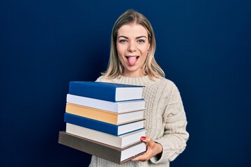 Beautiful caucasian woman holding a pile of books sticking tongue out happy with funny expression.