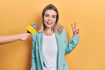 Beautiful caucasian woman being interviewed by reporter holding microphone smiling with happy face winking at the camera doing victory sign. number two.