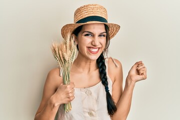 Young hispanic woman wearing summer hat holding spike wheat screaming proud, celebrating victory...