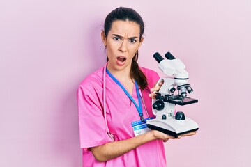Young brunette woman holding microscope in shock face, looking skeptical and sarcastic, surprised...
