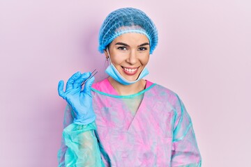 Young brunette woman wearing surgeon uniform holding tweezers looking positive and happy standing and smiling with a confident smile showing teeth