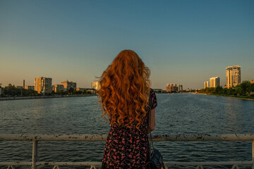 Sad redhead woman silhouette standing alone on a bridge in summertime sunset looking at urban view - 460545800