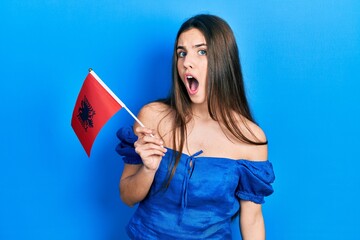 Young brunette teenager holding albania flag scared and amazed with open mouth for surprise, disbelief face