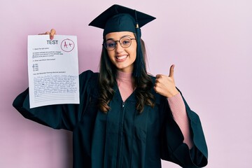 Young hispanic woman wearing graduation uniform holding passed exam smiling happy and positive, thumb up doing excellent and approval sign